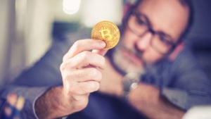 man in glasses holding a coin that has the Bitcoin (BTC-USD) logo