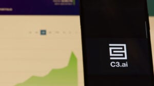 C3.ai (AI) logo on a smartphone with computer screen showing graph in background, symbolizing AI stock