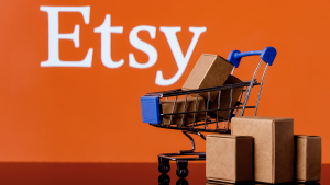 Etsy logo is over an orange background with a little shopping cart with packages in it. ETSY stock.