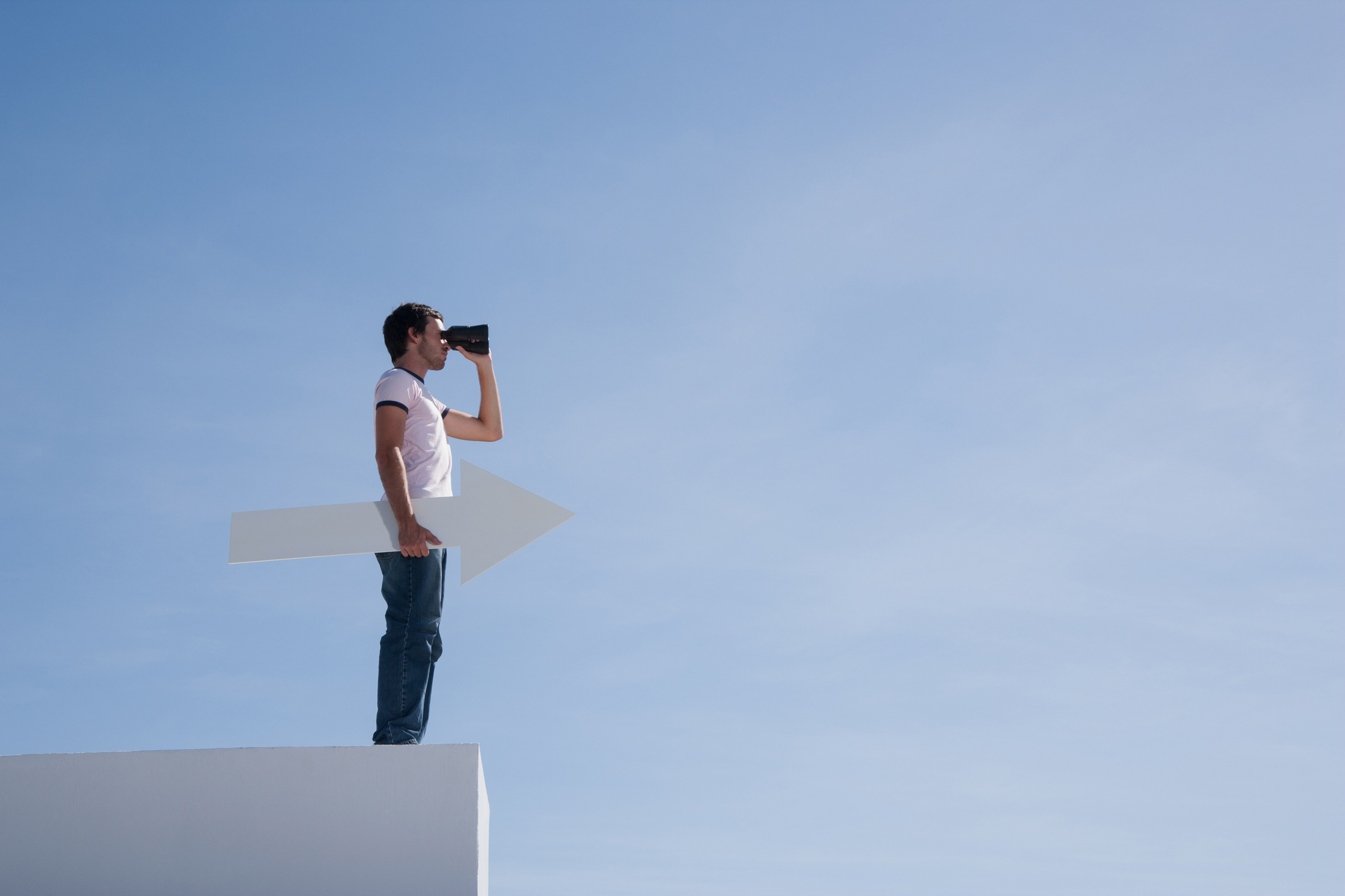 A person stands on a ledge holding binoculars and a paper cutout of an arrow.