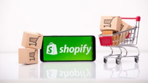 Shopify (SHOP) logo on a smartphone which is next to a miniature shopping cart and miniature cardboard boxes