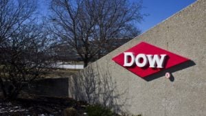 An image of a slanted concrete wall with a red diamond shaped sign on it with the text "Dow" in white, leafless trees, grass, and mulch around it, and a fence, a building, and a blue sky in the background.