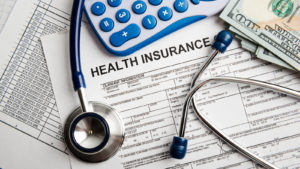 A health insurance claim form with a stethoscope, a calculator, and several hundred dollar bills resting on top.