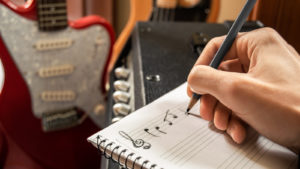 A photo of someone writing out music on staff paper with guitars in the background.