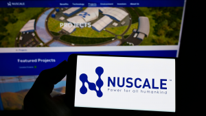 A hand in silhouette holds up a phone displaying the logo for Nuscale in front of a display showing the company's website.
