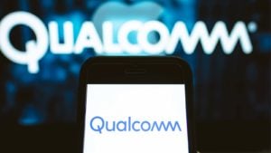An image of the top half of a black smartphone with a white screen displaying a blue "Qualcomm" logo, with a blurry white "Qualcomm" logo on a blue design in the background.