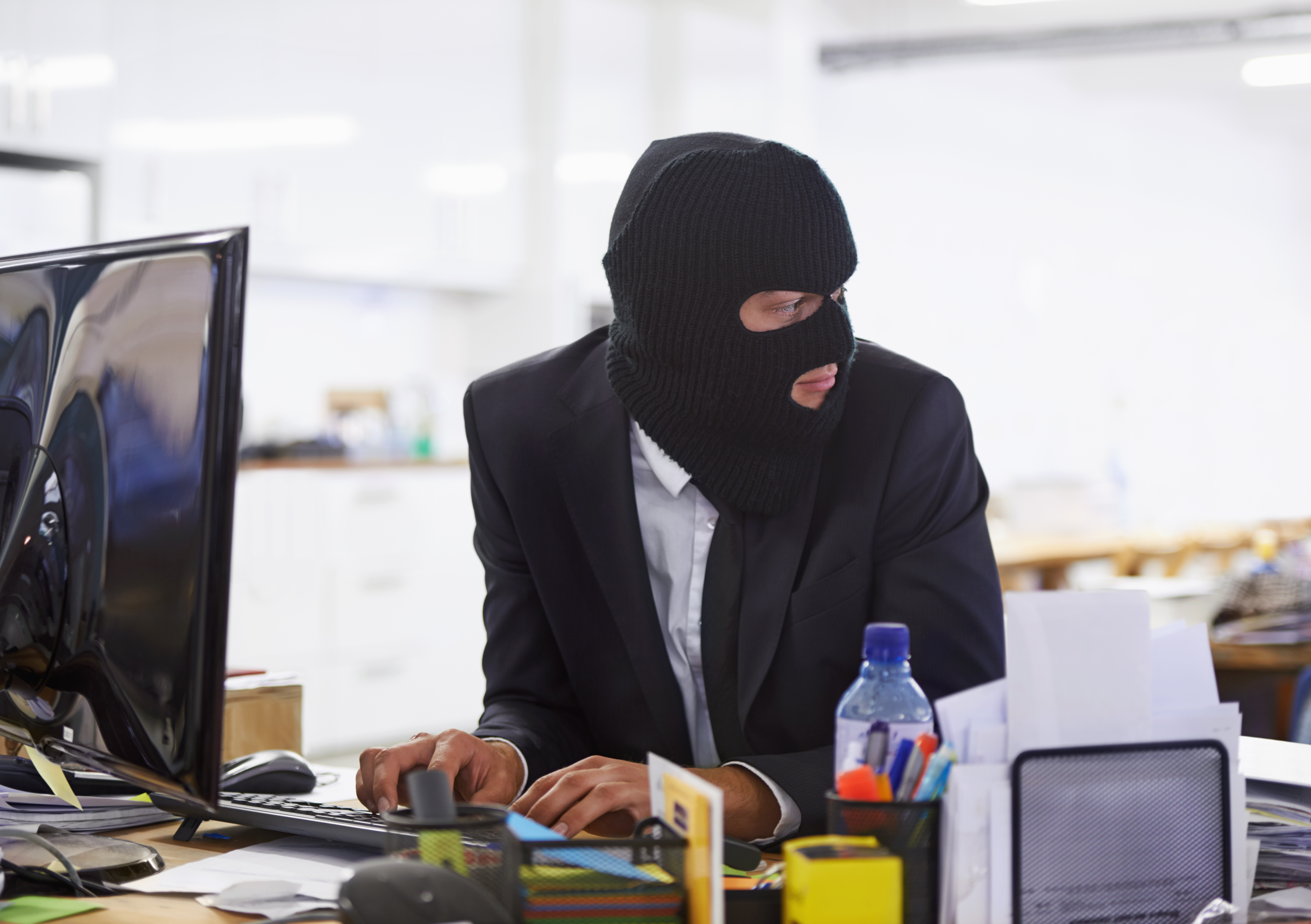 A person in a suit and ski mask typing on a computer in an office while looking over their shoulder.