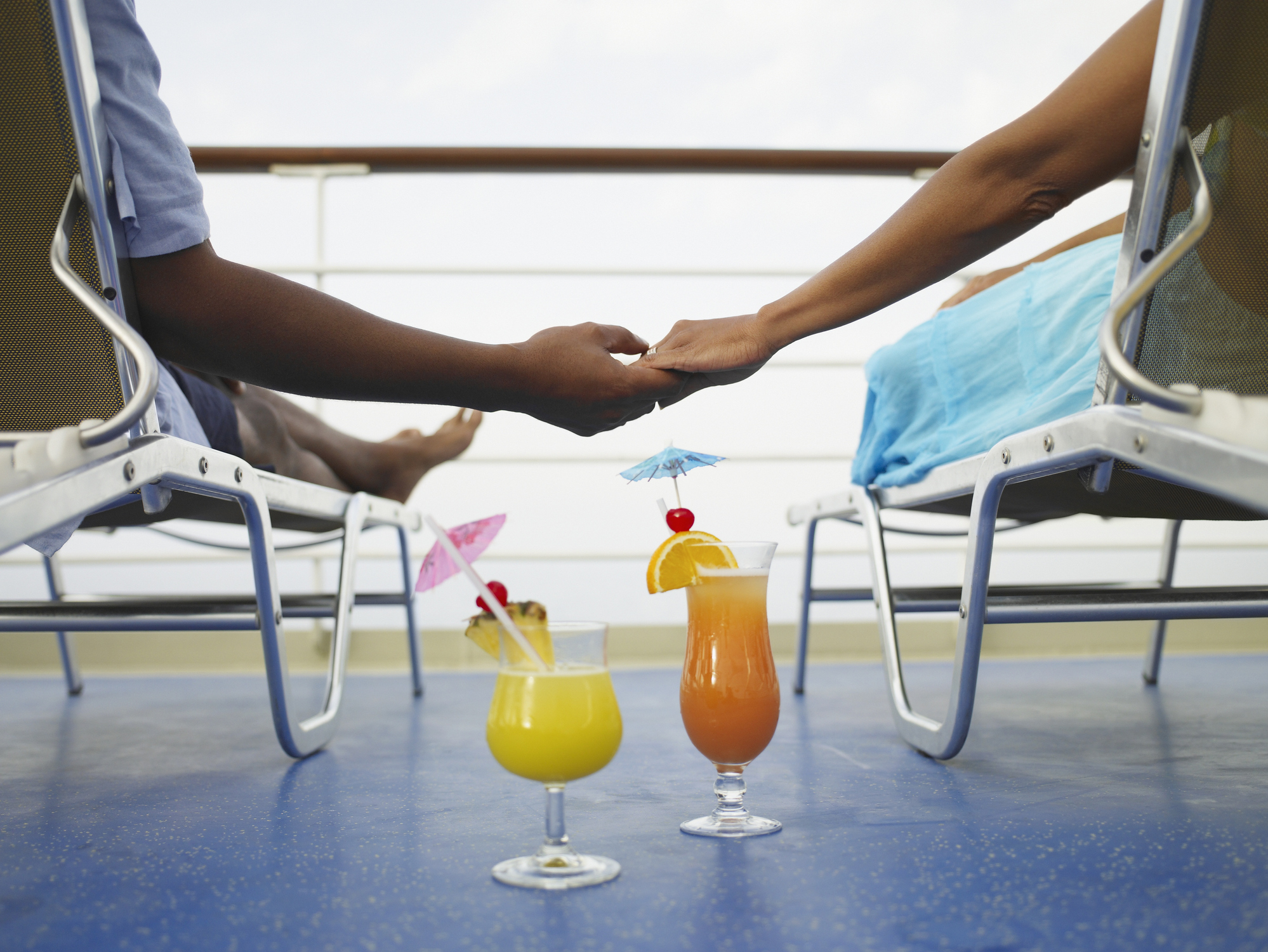 Two passengers holding hands on a cruise ship deck with tropical drinks between them.