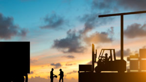 silhouettes of a forklift and driver as well as two workers by a semi truck backdropped by a sunset sky. represents the supply chain