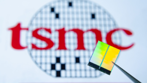 Close up photo of microchip (aka semiconductor chip, semiconductor device, Integrated Circuit) hold in tweezers with TSMC (TSM) logo on a background.