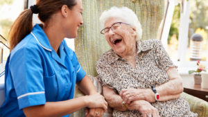 Senior Woman Sitting In Chair And Talking With Nurse In Retirement Home