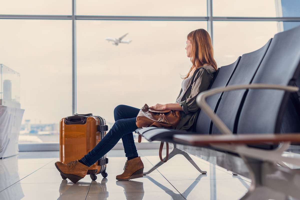 A person sitting at an airport waiting with their luggage as a plane takes off in the background.