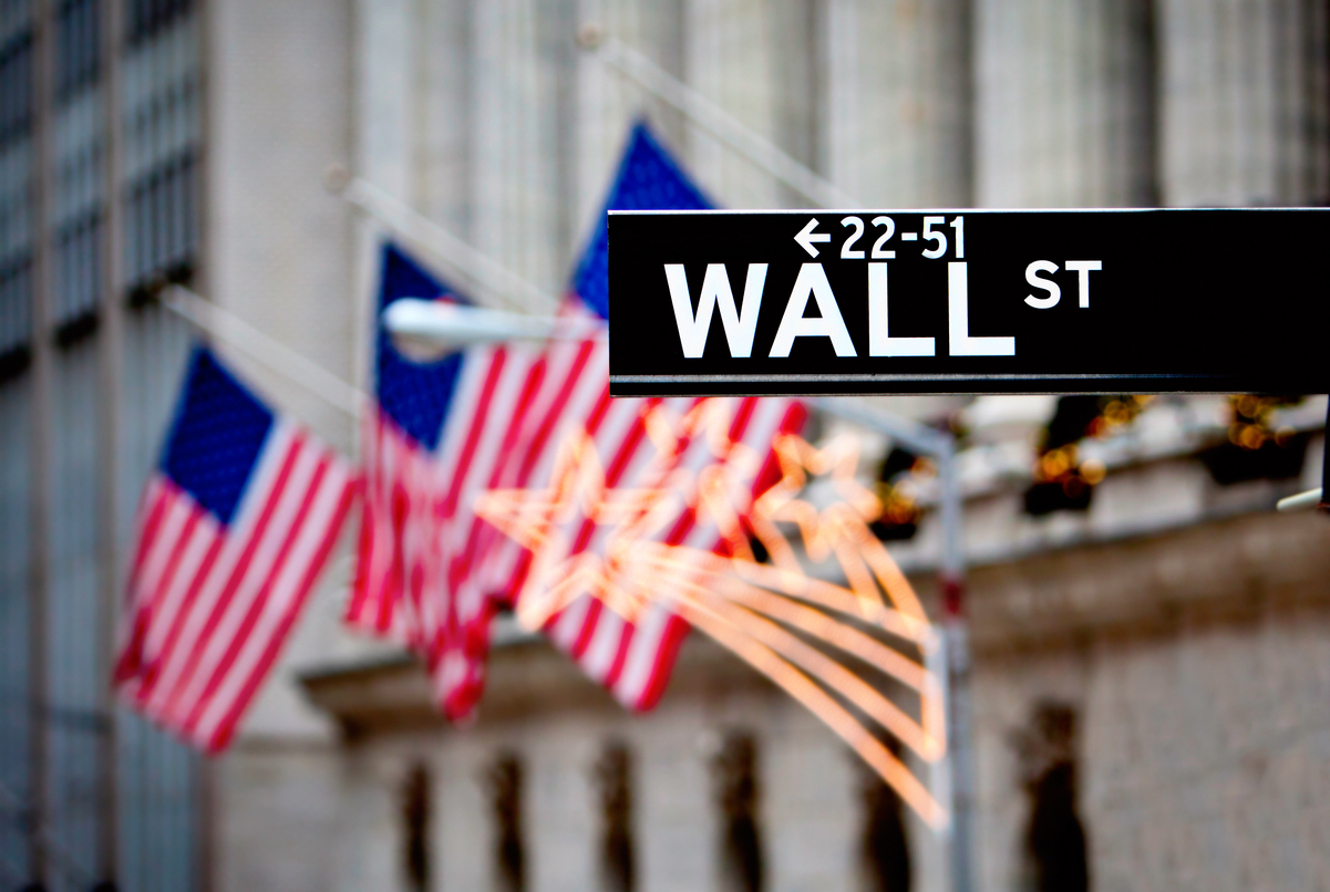 A street sign reading Wall St in front of a building with columns and American flags.