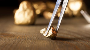 A photo of a gold nugget on a table, being picked up by tweezers, with more gold behind it. Stocks to Buy in March