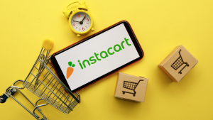 Instacart logo on a phone screen over a yellow background with a timer, boxes, and a shopping cart. Instacart IPO.