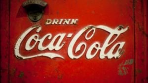 An image of an old red and rusted fridge door with a metal "Coca-Cola" logo handle and white embroidered text that reads "Drink" in a small font and "Coca-Cola" below in a very large font.