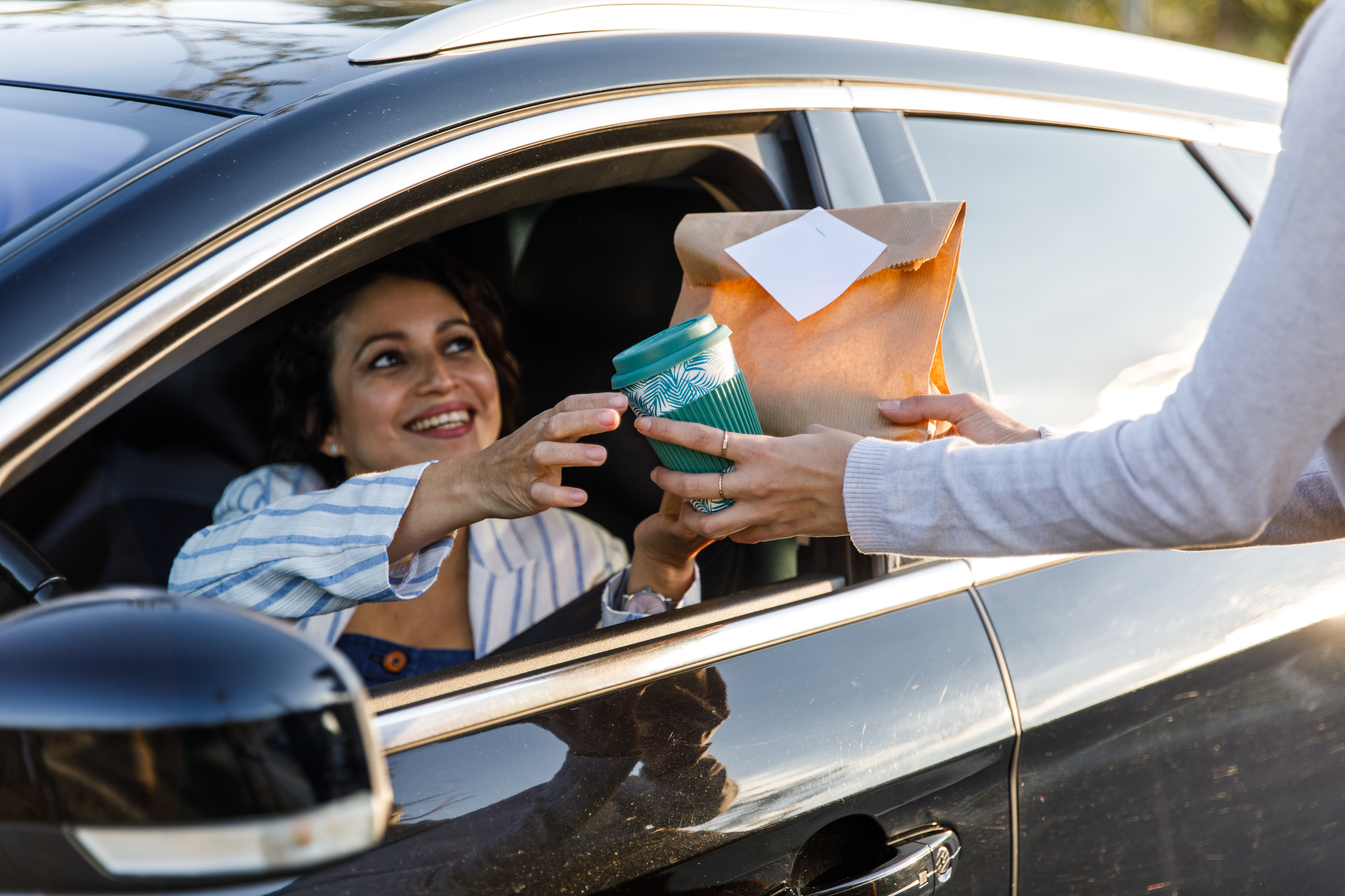 Smiling person receives coffee and a bag of snacks at a drive-thru window.
