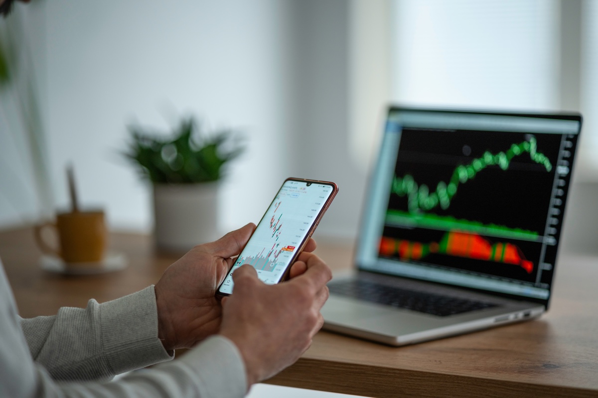 A person holding a phone with a graph on it while a computer displaying another graph sits on a nearby table.