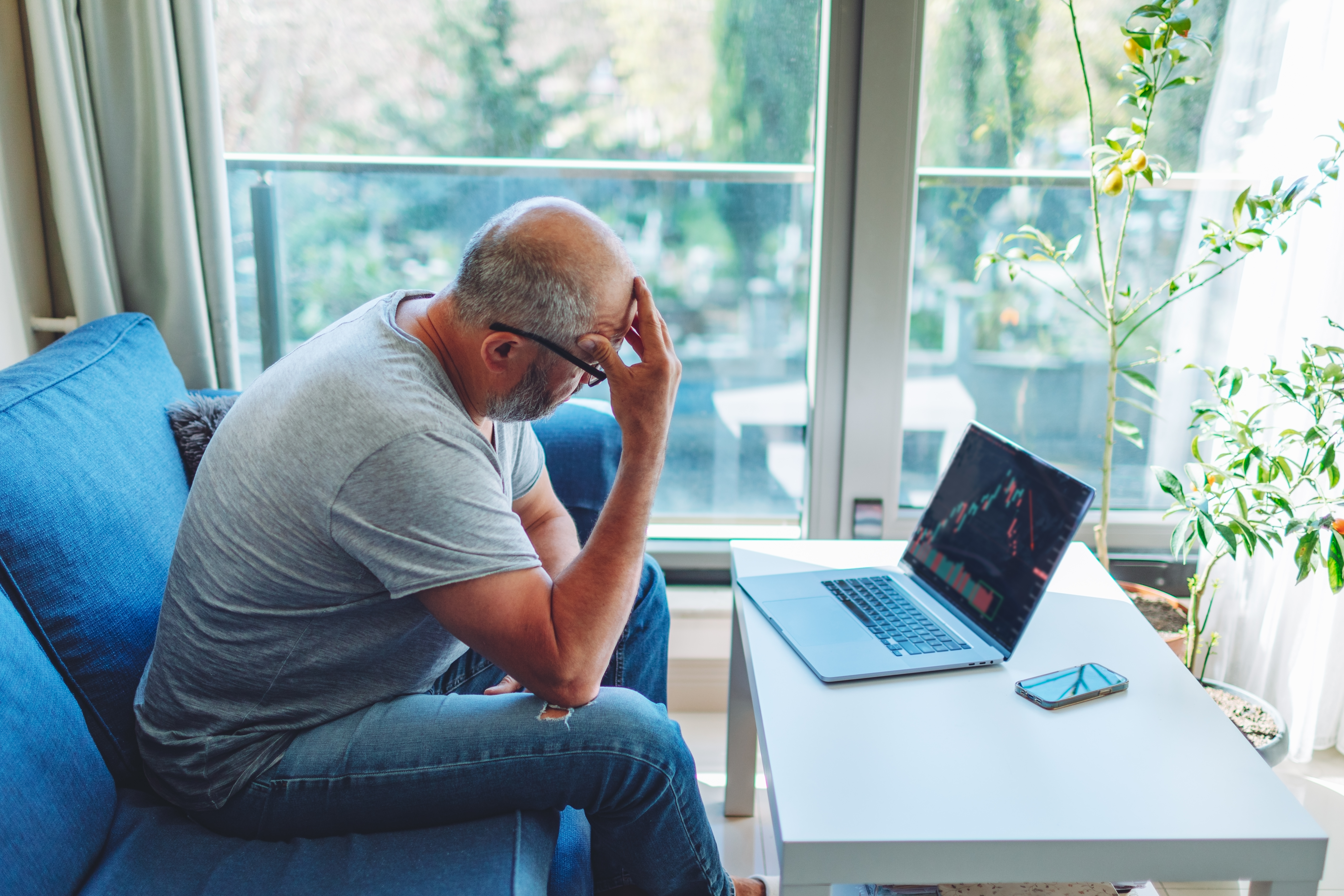 A person cringes while looking at laptop showing stock charts at home.