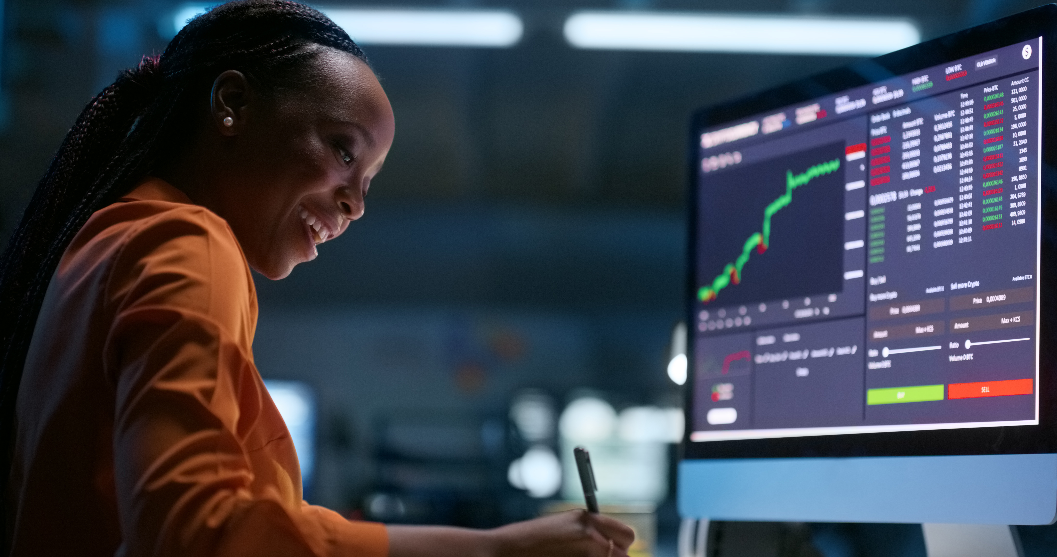 Person taking notes in front of a computer with stock charts on screen.